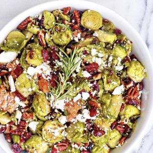 A round white bowl filled with roasted Roasted Brussels and Sausage, pecans, cranberries, and crumbled feta cheese. The dish is garnished with a sprig of fresh rosemary and displayed on a marble surface.