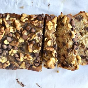 A sliced loaf of Gluten-Free Banana Bread topped with walnuts and chocolate chips on a white surface. The bread is light brown and looks moist, with a generous topping that adds texture and contrast.