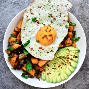 A white plate with a vibrant Sweet Potato Hash, mushrooms, and avocado slices, topped with a sunny-side-up egg. The dish is garnished with chopped herbs and red pepper flakes, set on a gray textured background.