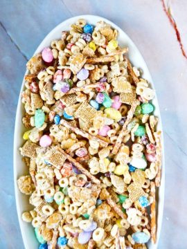 A white oval bowl filled with a colorful Easter Bunny Bait with various cereals, pastel-colored candy-coated chocolates, and pretzel sticks, set on a marble surface.