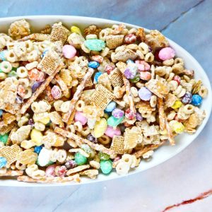 A white oval bowl filled with a colorful Easter Bunny Bait with various cereals, pastel-colored candy-coated chocolates, and pretzel sticks, set on a marble surface.
