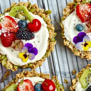 Delicious Granola Tarts topped with creamy yogurt, fresh berries, kiwi slices, chia seeds, and edible flowers, placed on a cooling rack over a marble surface.