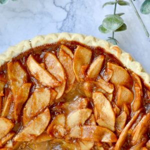 A freshly baked apple pie with a golden crust, topped with glazed apple slices, sits on a marble surface. Green leaves are visible beside the pie, adding a touch of nature to the scene.