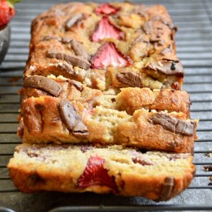 A loaf of Sweet Summery Strawberry Banana Bread is sliced and placed on a cooling rack. The bread has visible chunks of strawberries and chocolate pieces, set on a gray textured surface.