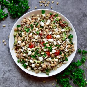 A bowl of Mediterranean Quinoa Salad with pine nuts, parsley, cherry tomatoes, feta cheese, and cucumber on a gray surface. Sprigs of fresh parsley are scattered around the bowl for garnish.