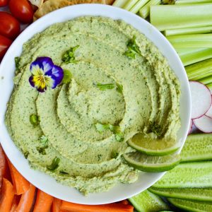 A platter with a bowl of creamy Cilantro Jalapeño Hummus garnished with lime slices and an edible flower, surrounded by sliced cucumber, radish, celery, carrot sticks, cherry tomatoes, crackers, and bell pepper slices on a marble surface.