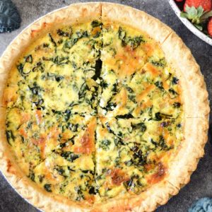 A baked Kale, Broccoli, And Cheddar Quiche with a flaky, golden crust sits in a round tin, several slices cut out to reveal a creamy, green-speckled filling. The quiche rests on a gray surface alongside fresh leafy greens.