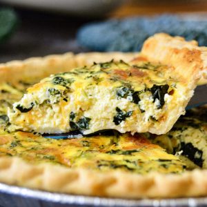A close-up of a freshly baked Kale, Broccoli, And Cheddar Quiche with a golden, flaky crust. A slice is being lifted, revealing a filling of eggs, cheese, and greens. The quiche sits in a foil pie dish, and a leafy vegetable is blurred in the background.