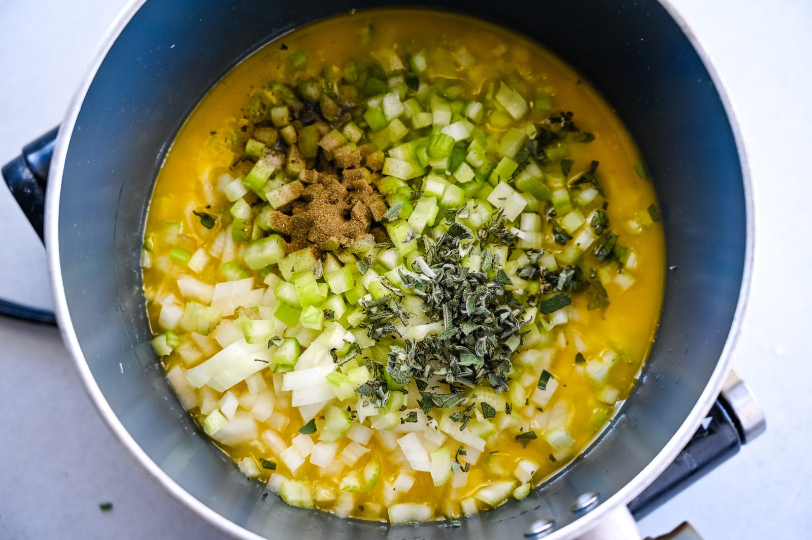 A pot on a stove contains a mixture of ingredients including chopped onions, diced celery, butter, and various herbs and spices. The mixture is being prepared for making Easy Stuffing.