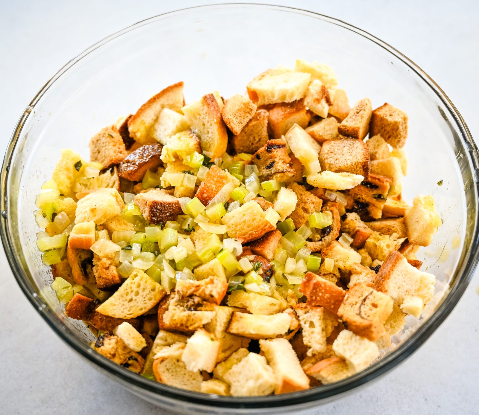 A large clear glass bowl filled with cubed bread pieces and diced celery, creating the base for a stuffing mix. The mixture appears fresh and undressed, ready for further preparation and seasoning.