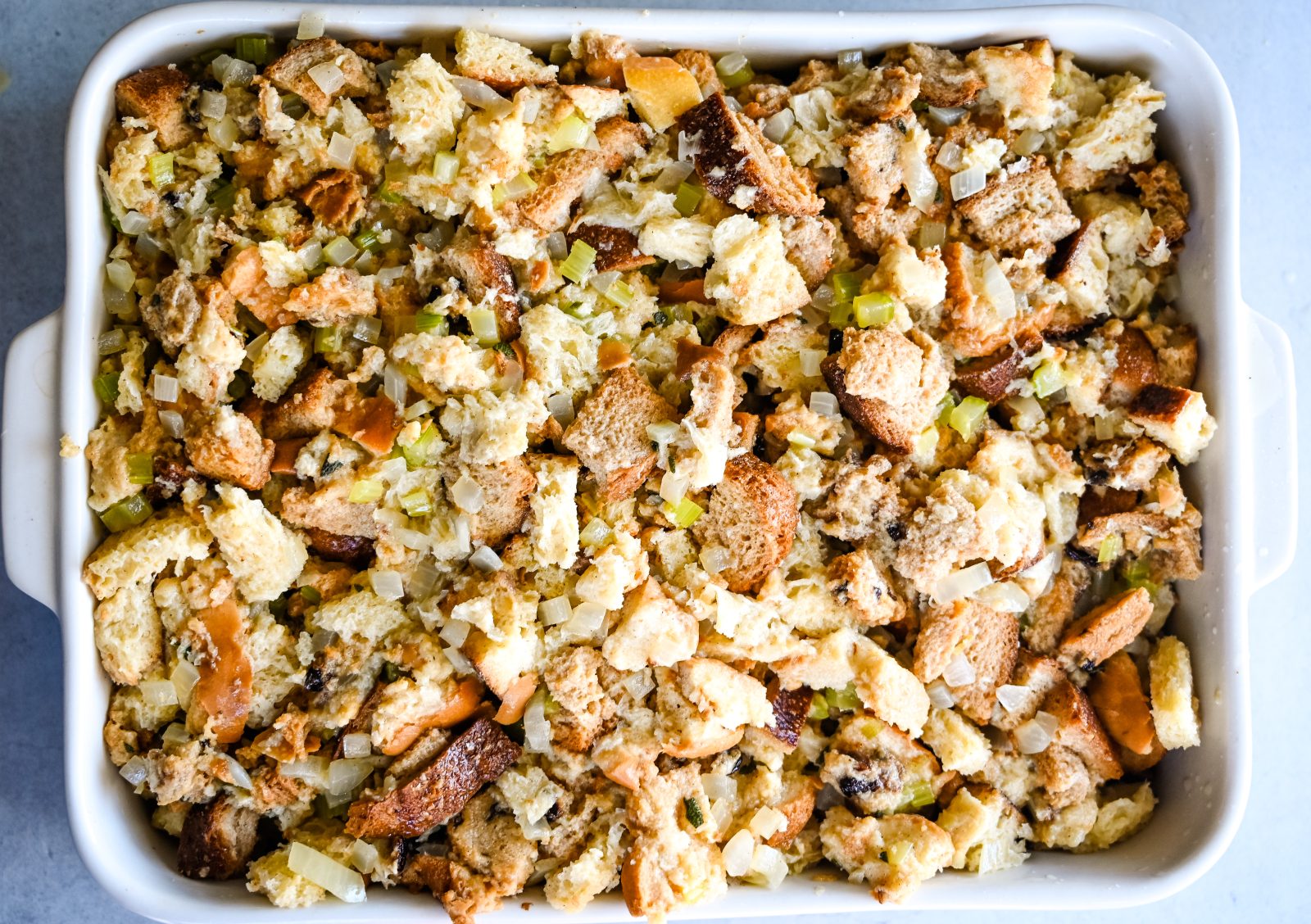 A white baking dish filled with unbaked Easy Stuffing made of cubed bread, chopped celery, onions, and herbs. The stuffing appears golden brown and slightly crispy on top, with bits of toasted bread and cooked vegetables clearly visible.