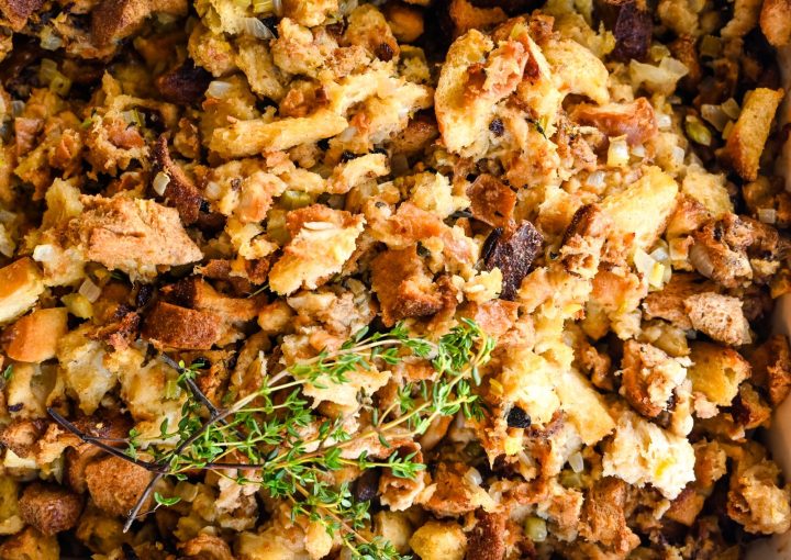 A close-up image of Easy Stuffing. The mixture includes various pieces of bread, some toasted, and appears to be seasoned with herbs and spices. A sprig of fresh thyme rests on top, adding a touch of green to the golden-brown dish.