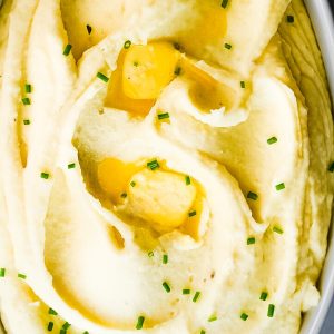 A dish filled with creamy mashed potatoes, topped with melted butter and garnished with finely chopped chives. The mashed potatoes appear smooth and velvety, with shallow dips holding pockets of melted butter, and the chives add a touch of green.