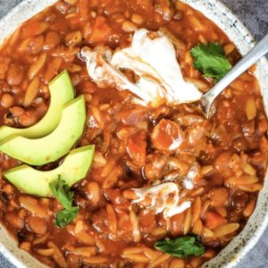 A bowl of hearty Instant Pot Vegan 10 Bean Taco Soup filled with beans, tomatoes, and orzo pasta, topped with sliced avocado, sour cream, and cilantro. A spoon is resting in the bowl on a textured gray surface.