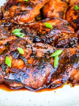 Grilled Easy Hawaiian Chicken glazed with a dark, sticky sauce are garnished with chopped green onions on a white plate. The chicken has a charred appearance, indicating it was cooked over high heat.