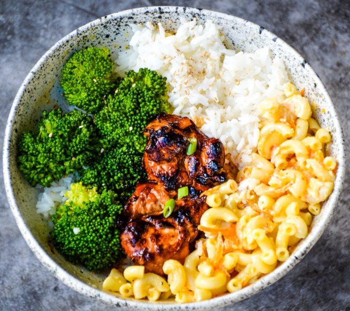 A bowl filled with white rice, Easy Hawaiian Chicken, steamed broccoli, and Healthier Hawaiian Macaroni Salad. The dish is presented against a textured gray background.