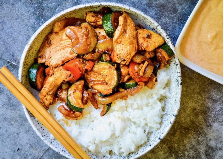 A bowl filled with sliced Hibachi Style Chicken & Veggies, including zucchini and red bell peppers, served over white rice. Chopsticks rest on the bowl's edge. A small dish with a creamy sauce is placed beside the bowl.