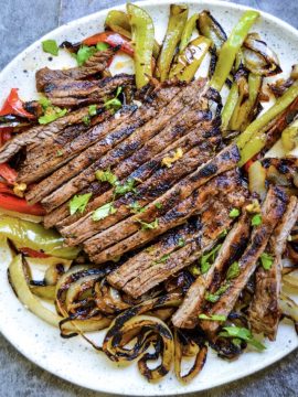 A plate of Restaurant-Style Steak Fajitas with strips of grilled beef, red and green bell peppers, and onions. Garnished with lime wedges and cilantro, set on a rustic gray surface.