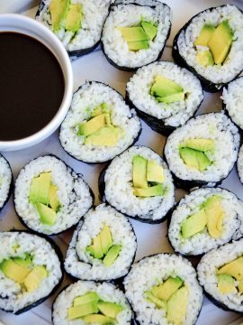 A plate of sliced Avocado Sushi Rolls filled with rice and avocado, served with a small dish of soy sauce on the side. The sushi rolls are arranged in a circular pattern on a textured surface.