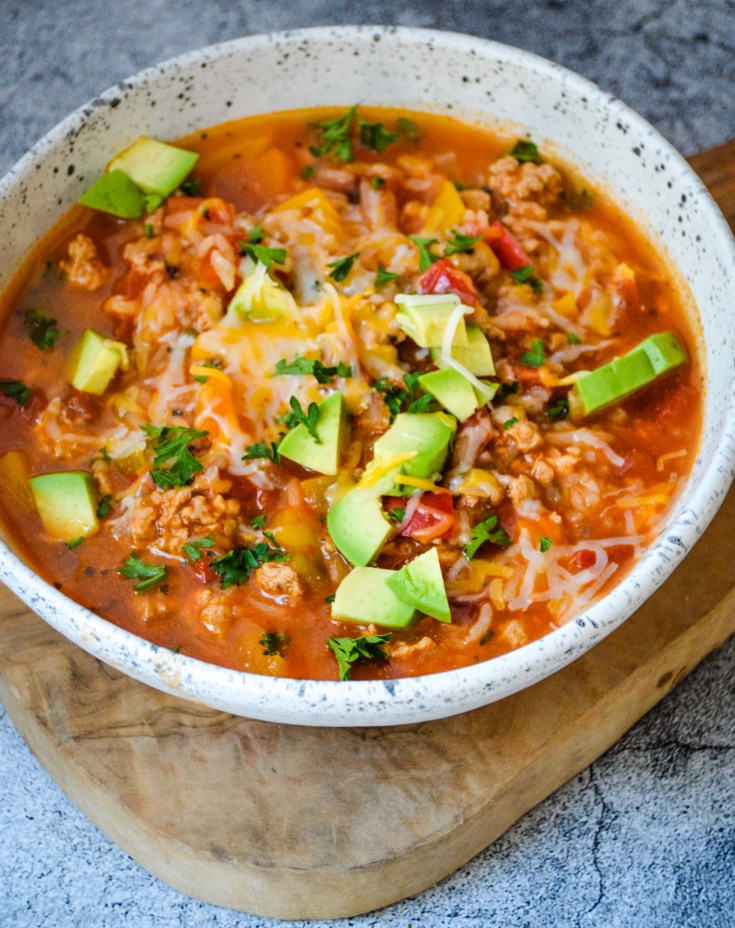 A savory bowl of Stuffed Pepper Soup featuring a blend of ground meat, tomatoes, diced avocados, shredded cheese, and chopped herbs, served in a white speckled bowl placed on a wooden cutting board. The dish is colorful and garnished with fresh parsley.