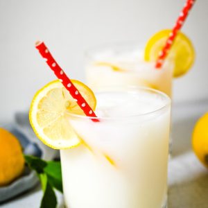 Two glasses filled with refreshing Creamy Lemonade, each garnished with a lemon slice on the rim and red polka-dot straws. Fresh lemons and green leaves are slightly visible in the background.