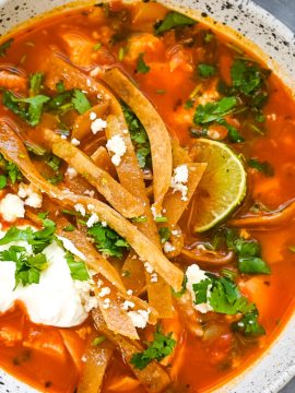 A bowl of Easy Chicken Tortilla Soup, adorned with crispy tortilla strips, a lime wedge, fresh cilantro, and a dollop of sour cream. The rich, red broth is filled with chunks of chicken and vegetables. A gray napkin is partially visible beside the bowl.