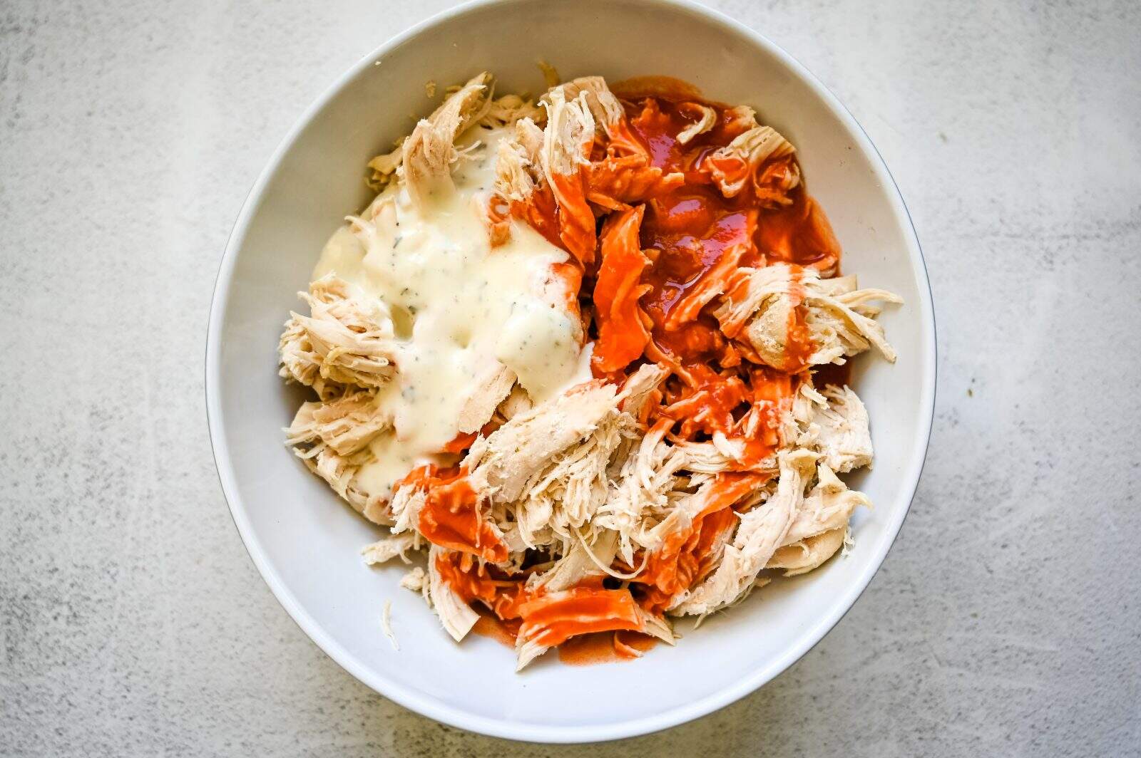 A bowl filled with shredded chicken, topped with sections of orange hot sauce and a creamy white sauce. The white background contrasts with the vibrant colors of the sauces.