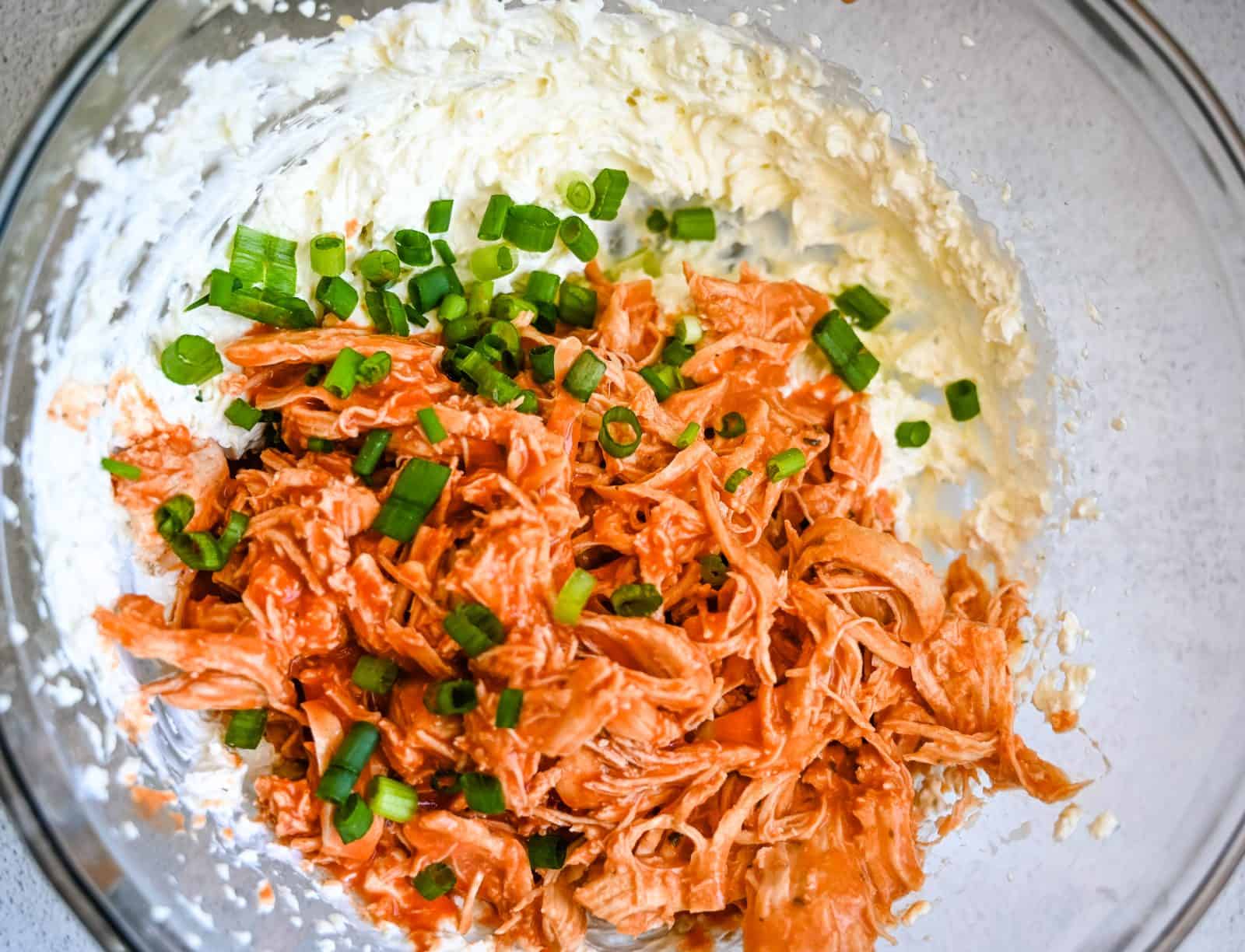 A close-up view of creamy, whipped cream cheese in a glass bowl, topped with shredded buffalo chicken and green onions. The texture is light and fluffy with a pale yellow color, and the bowl is placed on a light gray surface.