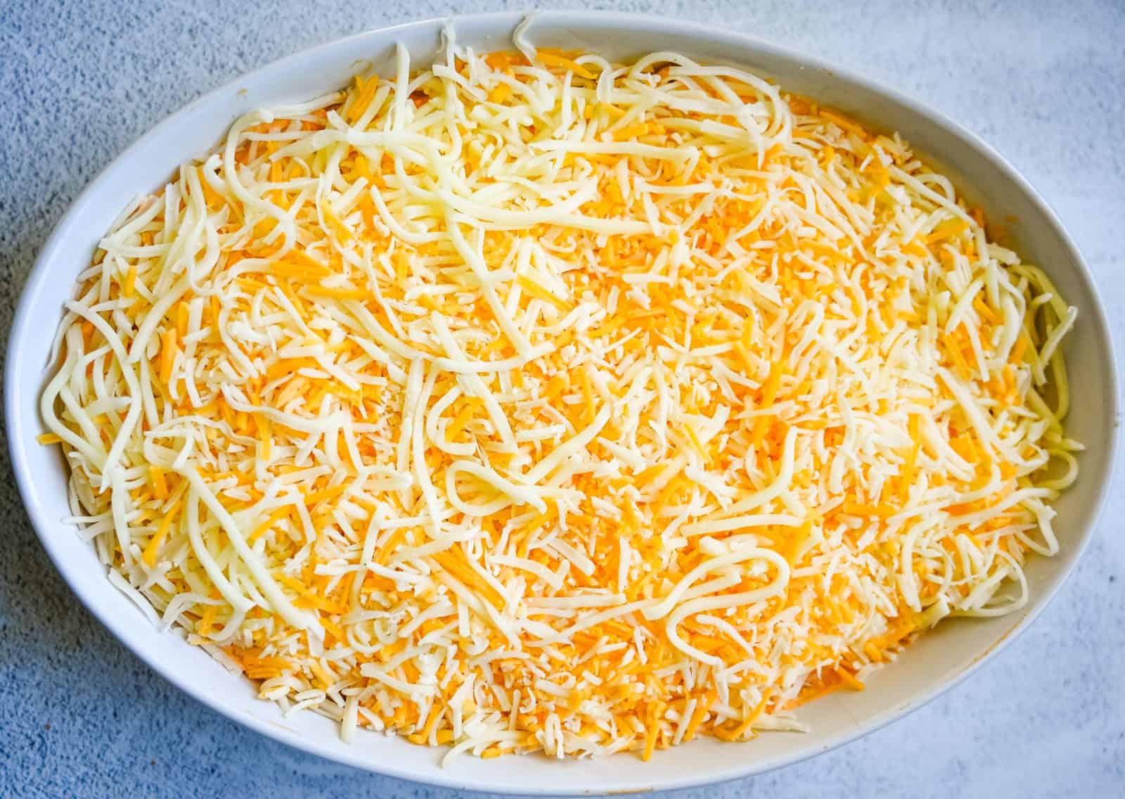 Oval white dish filled with creamy, orange buffalo chicken dip, that has been topped with a shredded blend. The background surface is light-colored and textured.