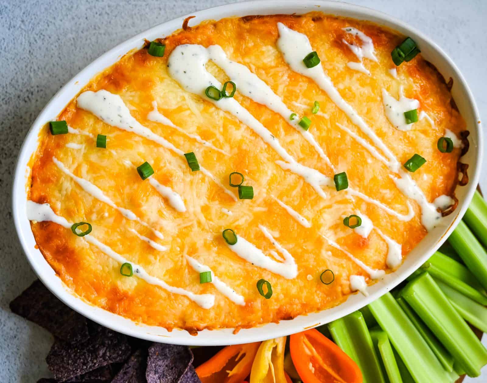 Baked Buffalo Chicken Dip garnished with chopped green onions and drizzled with white sauce, served in an oval dish. Beside the dip are celery sticks, sweet mini bell peppers, and blue tortilla chips arranged on a white surface.