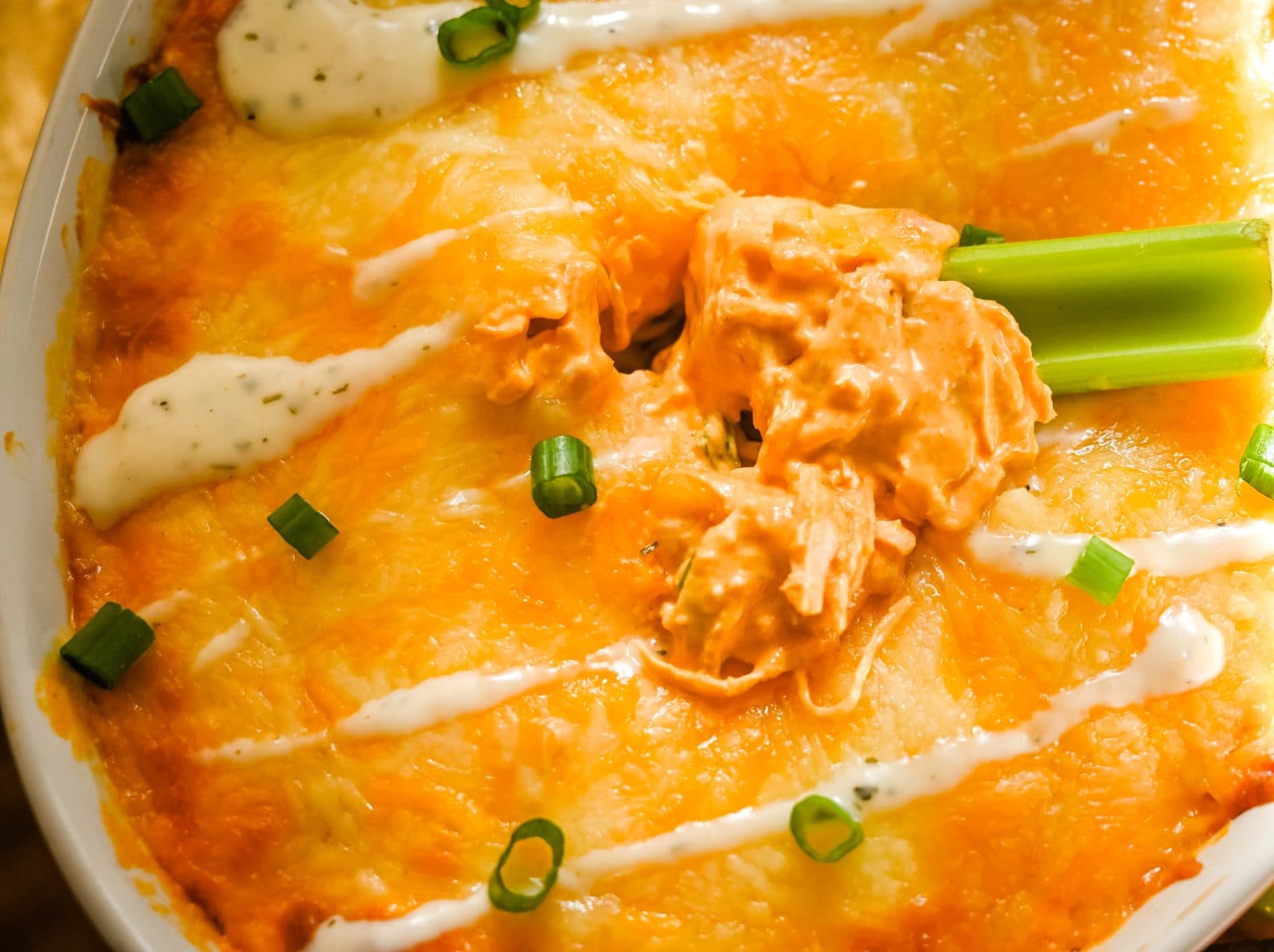 Close-up of a cheesy, oven-baked Buffalo Chicken Dip topped with chopped green onions and a piece of celery. The golden, bubbly cheese covers a creamy, orange-tinted layer beneath. Drizzled with creamy ranch dressing, adding contrast to the dish.