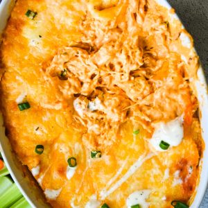 A close-up of a baked dish filled with a cheesy buffalo chicken dip. The top is golden brown with melted cheese, some green onions sprinkled on top, and a few dollops of creamy dip. A portion of the dip has been scooped out, revealing the shredded chicken underneath.