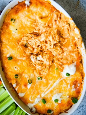 A close-up of a baked dish filled with a cheesy buffalo chicken dip. The top is golden brown with melted cheese, some green onions sprinkled on top, and a few dollops of creamy dip. A portion of the dip has been scooped out, revealing the shredded chicken underneath.