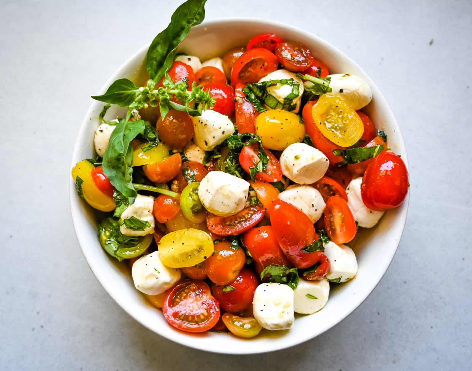 A bowl of colorful Cherry Tomato Caprese Salad featuring cherry tomatoes, mozzarella balls, and basil leaves, all lightly dressed. The tomatoes come in red, yellow, and green, contrasting with the white mozzarella and green basil for a vibrant, fresh presentation.
