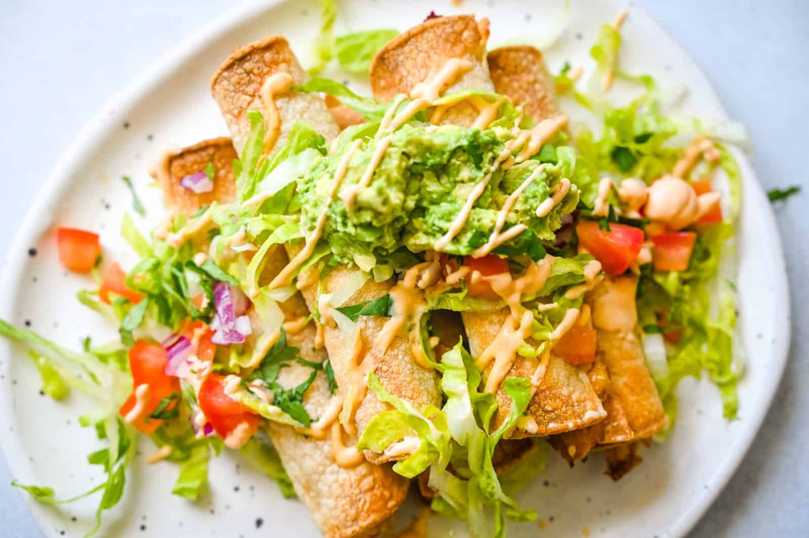 A close-up image of a plate of crispy Chicken Taquitos topped with chopped lettuce, diced tomatoes, guacamole, and a drizzle of Chipotle Crema. The dish is colorful and appetizing, capturing the details of the fresh ingredients.