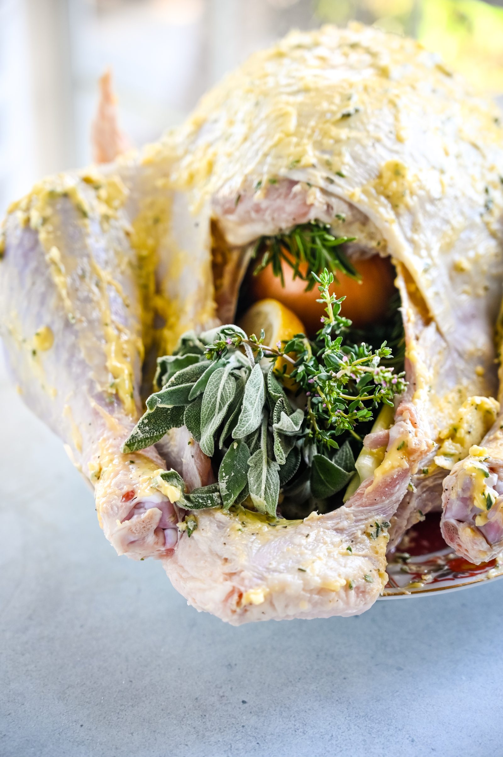 A raw, uncooked turkey with its cavity filled with fresh herbs, garlic, and orange slices. The turkey is seasoned with an herb butter and is ready for roasting, placed on a light-colored surface.