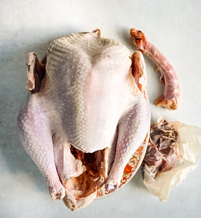 A raw, whole turkey placed on a plate with its neck and giblets beside it on a light-colored surface. The turkey is prepped and ready for further preparation or cooking.