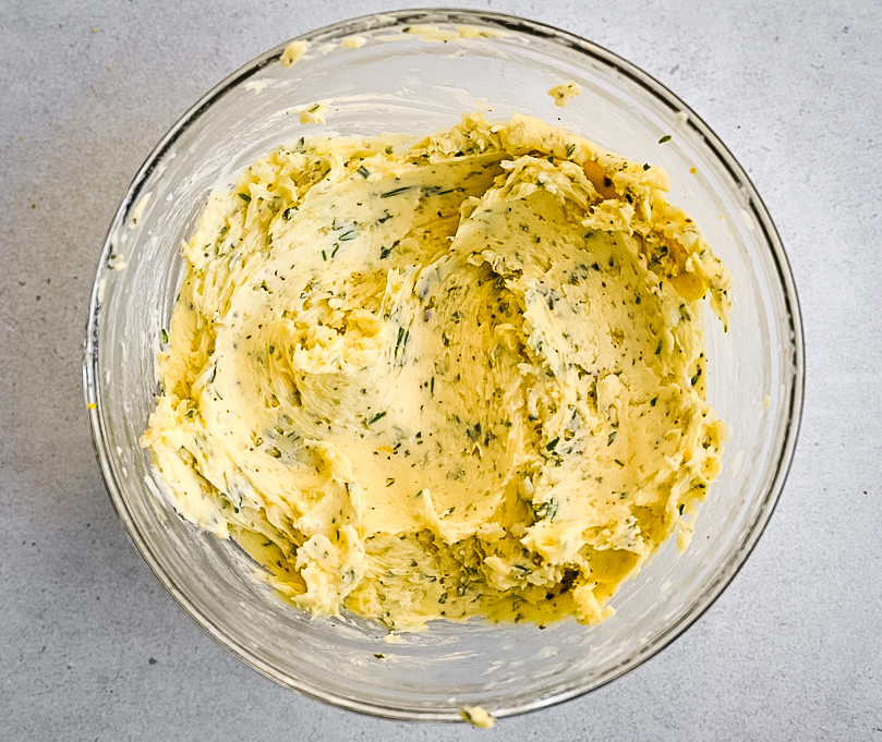 A clear glass bowl containing a mixture of softened butter blended with finely chopped herbs, placed on a light gray surface. The butter is smooth and creamy, with visible green herb flecks scattered throughout.