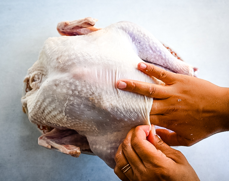 Two hands are loosening the skin of a raw turkey, preparing it for seasoning or stuffing. The turkey is placed on a light surface, and one hand is gently lifting the skin while the other is holding the chicken steady.