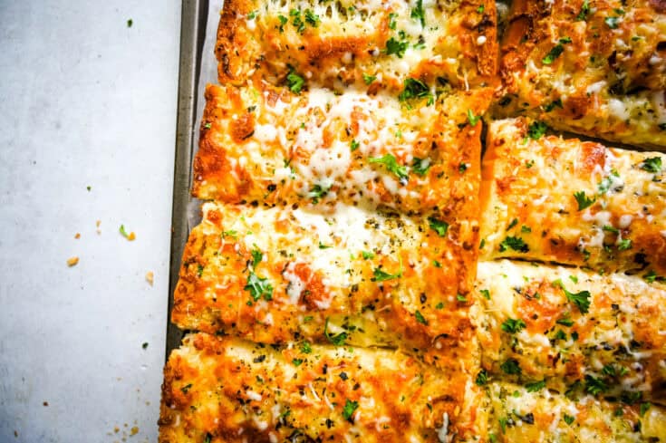 Close-up of slices of cheesy garlic bread topped with melted cheese and garnished with chopped parsley. The bread has a golden-brown crust, and some crumbs are visible on the white paper background, highlighting the texture and freshness of the slices.