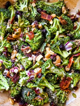 A wooden bowl filled with a vibrant broccoli salad. The salad contains chopped broccoli florets, crispy bacon pieces, dried cranberries, red onion, sunflower seeds, and peanuts, all mixed with a creamy dressing.