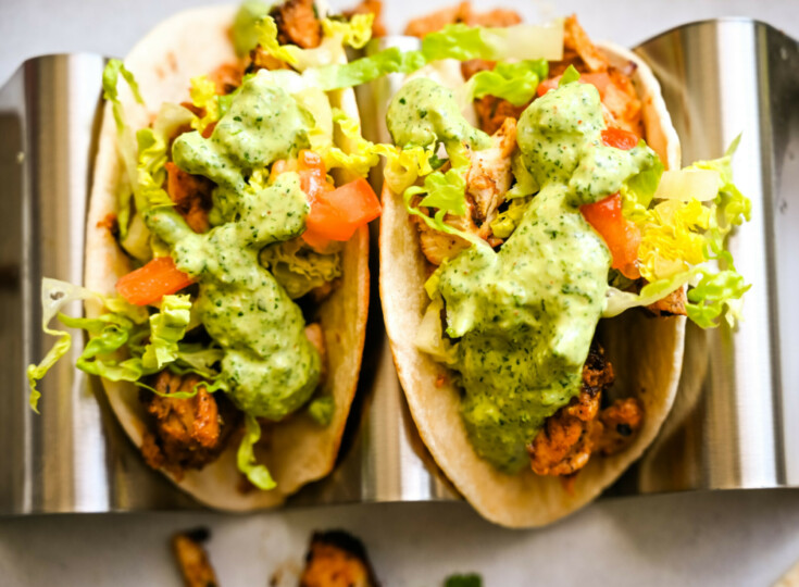 Close-up of Chicken Tacos with Creamy Cilantro Sauce in a metal holder. The taco is filled with grilled chicken, chopped lettuce, diced tomatoes, and topped with a generous serving of creamy cilantro sauce. The bright colors and fresh ingredients make it look appetizing.