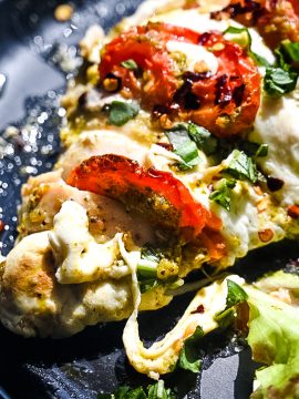 Close-up of Hasselback Caprese Chicken featuring baked chicken breast topped with melted mozzarella cheese, sliced tomatoes, fresh basil, and a sprinkle of red pepper flakes on a dark plate. Fresh mixed greens are visible in the corner for a side serving.