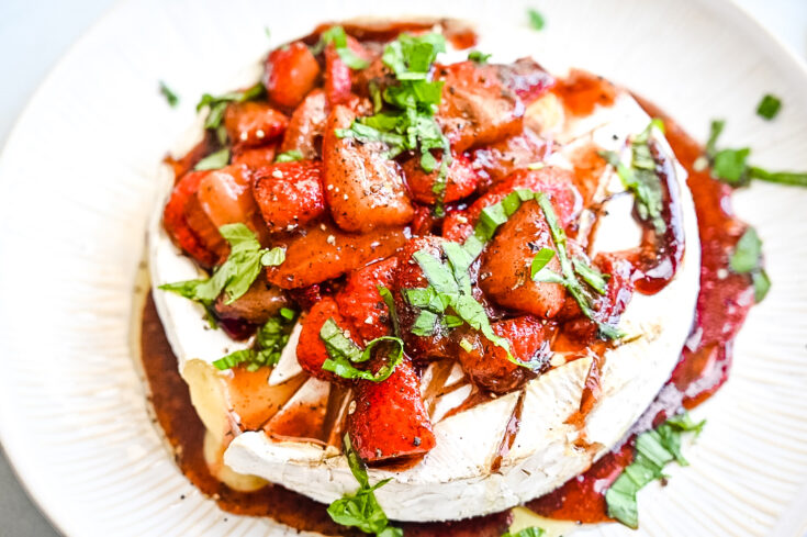 Close-up of a baked brie topped with a vibrant mixture of roasted strawberries, a drizzle of balsamic glaze, and sprinkled with fresh chopped basil. The dish is presented on a white plate, accentuating the rich colors and textures of the toppings and cheese.