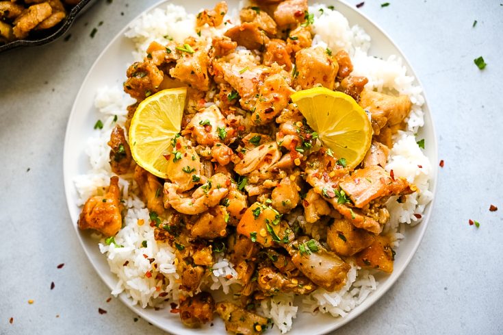 A close-up of a plate of flavorful dish featuring Honey Garlic Chicken Bites garnished with red pepper flakes and chopped herbs, served over a bed of white rice. Two lemon slices are placed on top for added acid and freshness.