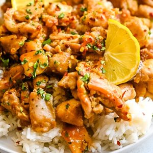 A close-up of a plate of flavorful dish featuring Honey Garlic Chicken Bites garnished with red pepper flakes and chopped herbs, served over a bed of white rice. Two lemon slices are placed on top for added acid and freshness.