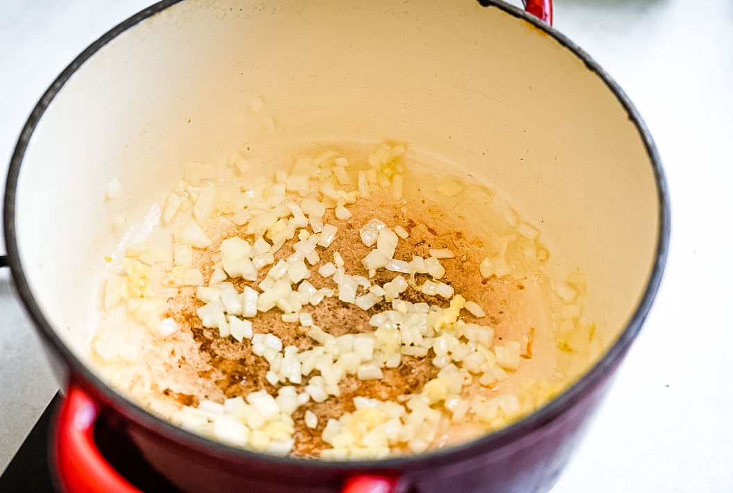 Chopped onions are sautéing in a large red pot, creating a base for cooking. The onions are becoming translucent, indicating they are being cooked over medium heat. The inside of the pot shows light browning, adding to the flavor development.