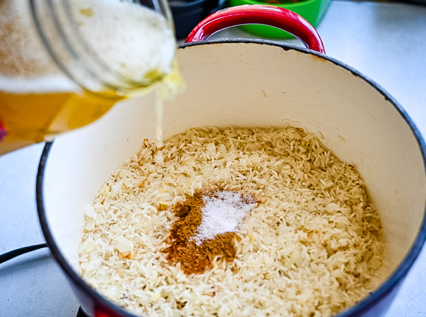 A close-up of a blue and red pot containing uncooked rice, with ground spices, and salt on top. A jar of broth is being poured into the pot, with visible foam from the liquid. The cooking scene is set on a kitchen counter.