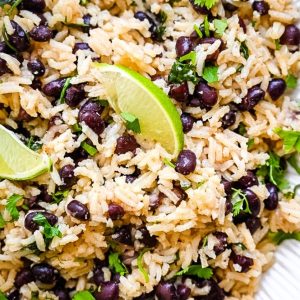 A plate of Black Beans and Rice, garnished with fresh cilantro and lime wedges. The dish appears to be seasoned with various herbs and spices, giving it a flavorful and visually appealing presentation.