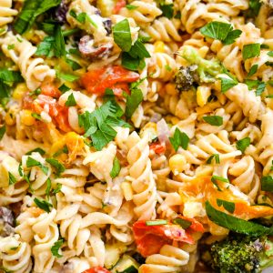 Close-up of a colorful hummus pasta salad featuring rotini pasta, diced tomatoes, corn kernels, broccoli florets, and black olives. The dish is garnished with chopped parsley and appears to be dressed in a creamy sauce.